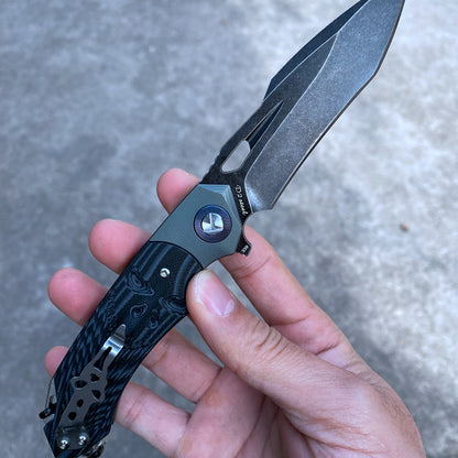 Hand holding a D2 steel survival folding knife with a G10 handle and titanium alloy elements, showcasing the blade and intricate craftsmanship.