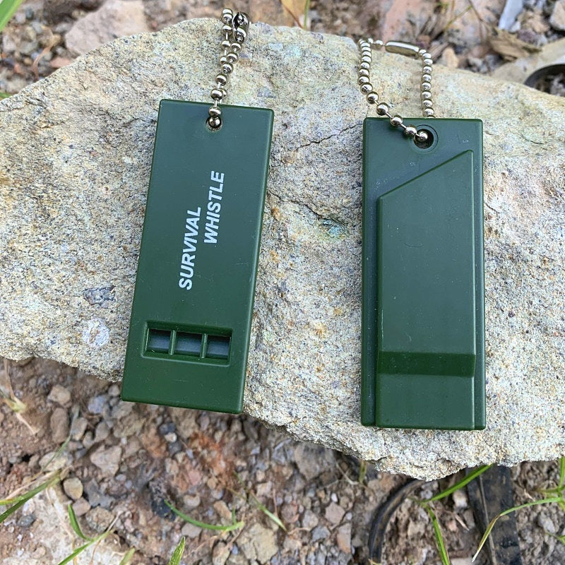 Two dark green high decibel survival whistles with chains, placed on a rock, showing front and back views.