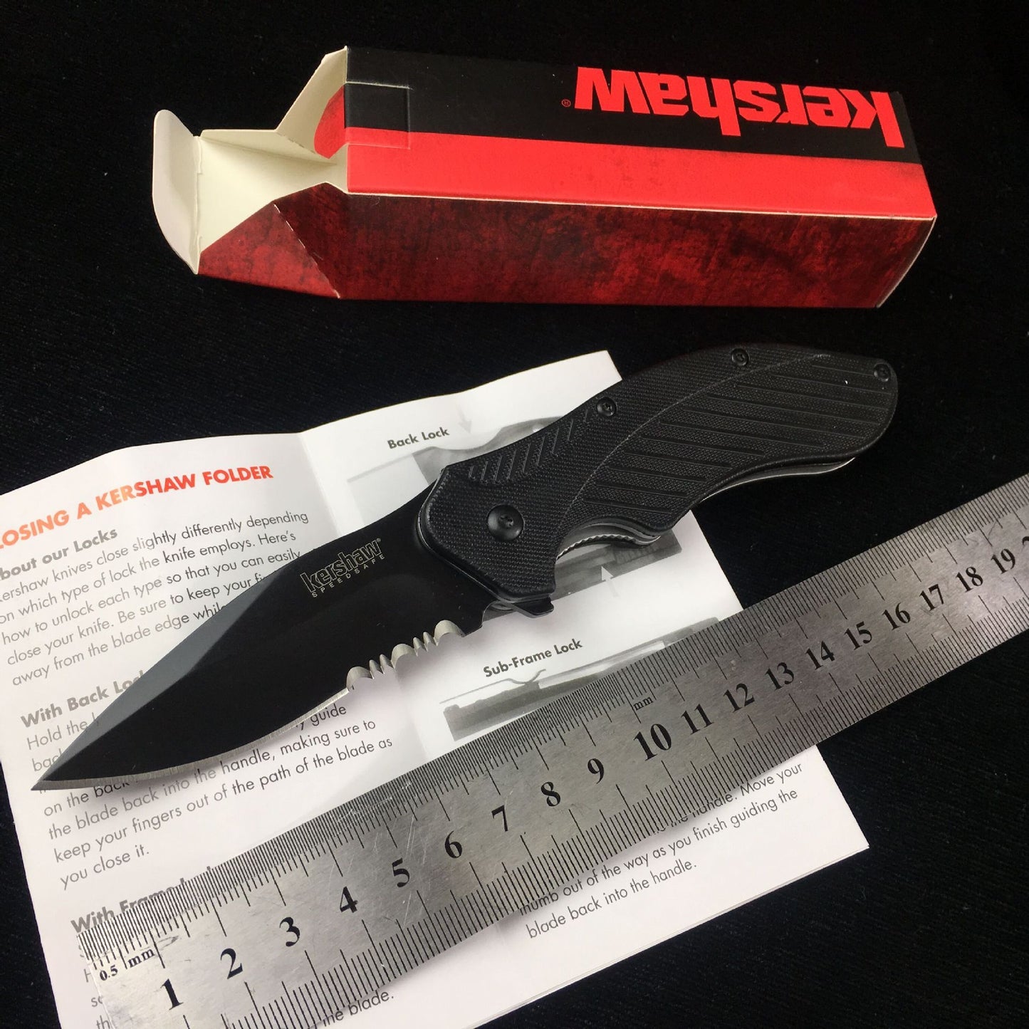 Kershaw survival folding knife with black handle and exposed blade, placed next to an open box and a ruler for size reference.